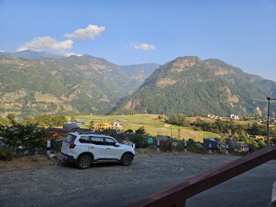 A white car parked on a hillside road above terraced fields, with forested mountains beyond.