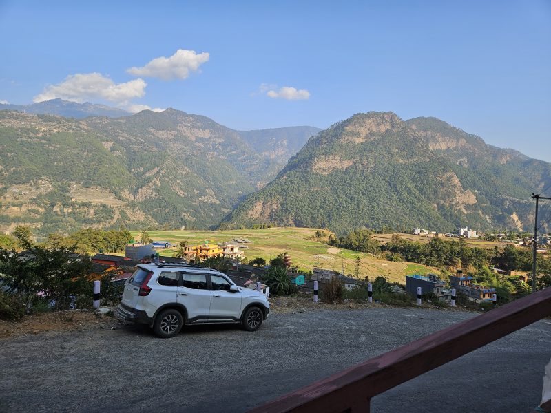 A white car parked on a hillside road above terraced fields, with forested mountains beyond.