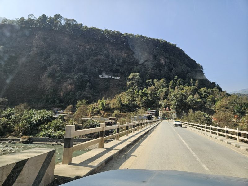 A road crossing a bridge towards a forested mountainside, seen through a car windscreen.